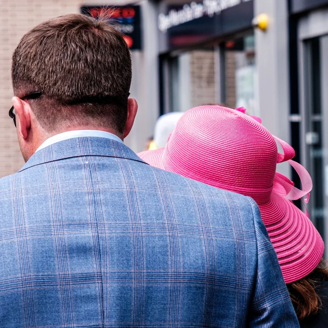 Back view of a man in a blue checked suit jacket and a woman in a bright pink hat, stylishly dressed for a British summer racing event.