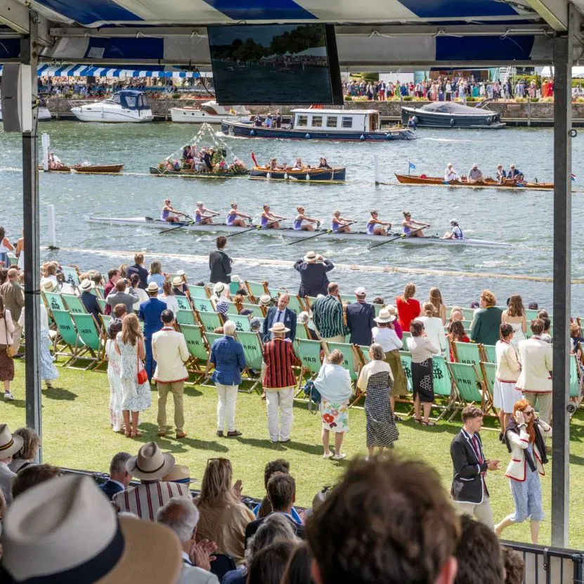 Spectators in blazers and straw hats watching rowing crews compete on the Thames at Henley Royal Regatta, with boats and crowds lining the opposite riverbank.