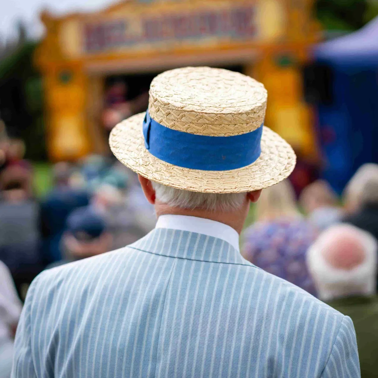 Back view of a man in a blue pinstripe suit and straw boater hat with blue ribbon, dressed for the Kentucky Derby – classic men’s Derby hat style.