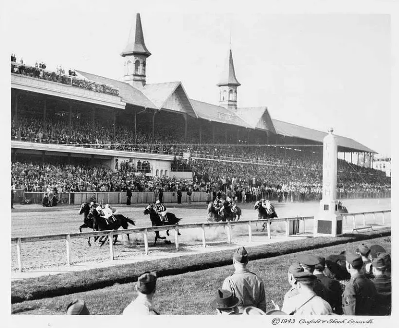 Historic black and white photo of horse racing at Churchill Downs, with packed grandstands and the iconic Twin Spires during the Kentucky Derby.