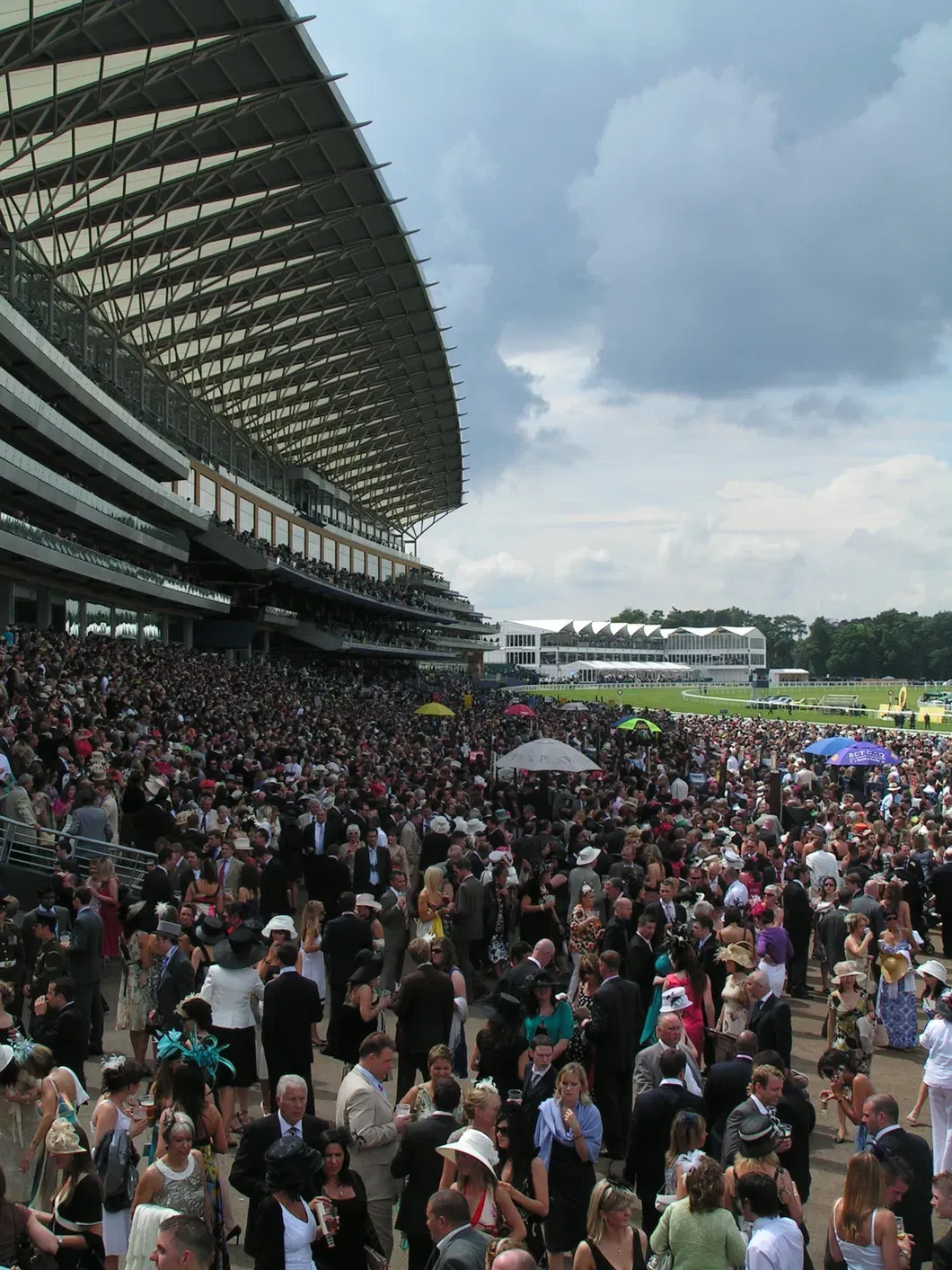 Large crowd in formal attire at Royal Ascot racecourse, with grandstand and dramatic sky during the British social season.