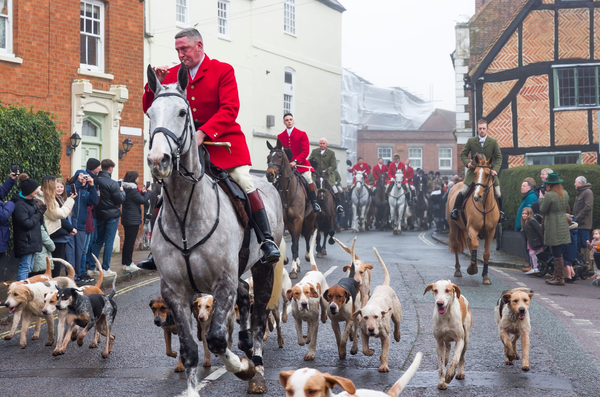 Boxing Day hunt meet in an English village street with hounds, mounted field in red coats, and spectators lining the road