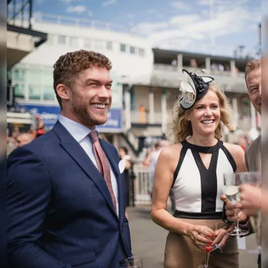 Gentlemen and ladies dressed in classic Kentucky Derby style, featuring a tailored navy suit, elegant fascinator, and champagne flutes at a race day event.