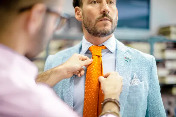 Tailor adjusting an orange tie on a man wearing a light blue linen jacket and pocket square – Derby Day style and linen jacket fit for the Kentucky Derby.