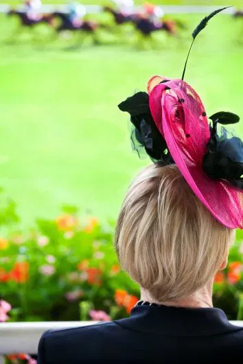 Woman in a bright pink bespoke Derby hat watching horse racing from the grandstand – women’s hat style at the Kentucky Derby.